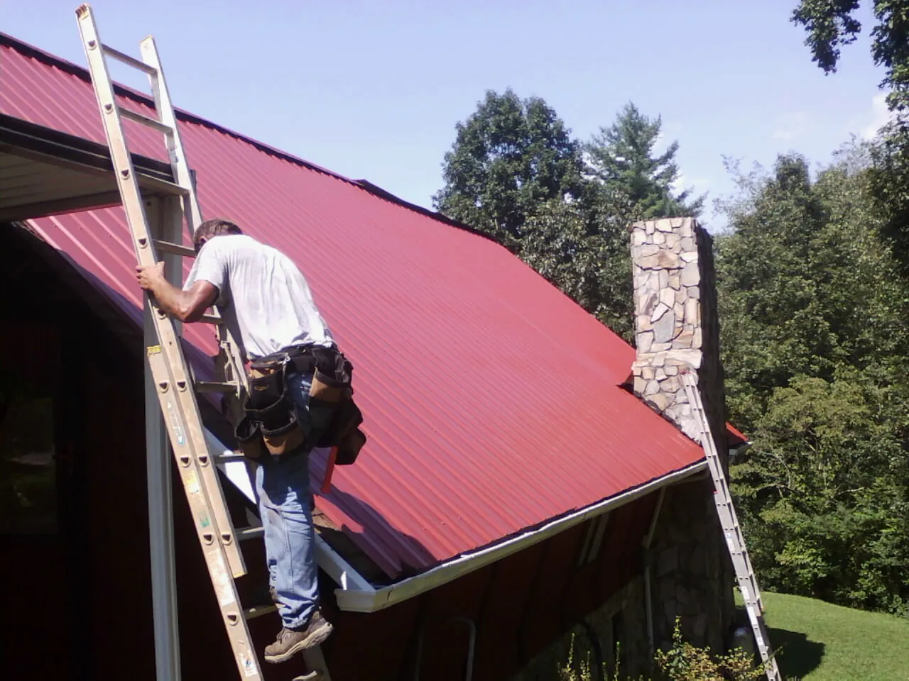 Roofer on a ladder beside a red metal roof and stone chimney — Soffit & Fascia Repair by Roofing Brothers in Pacolet Mills